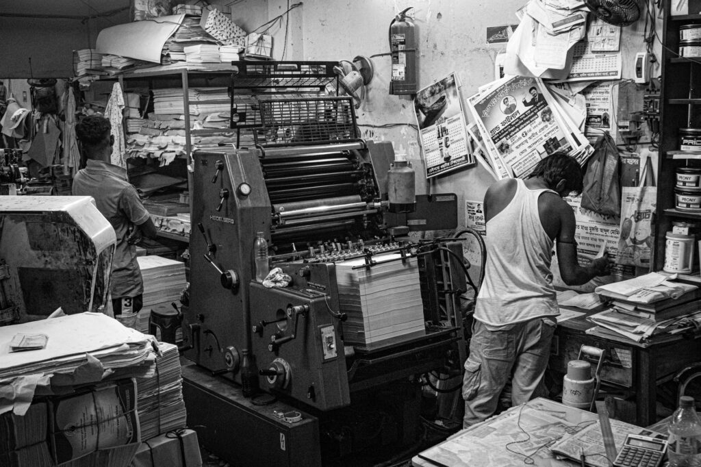 Black and white image of a bustling print shop with workers operating a printing press.